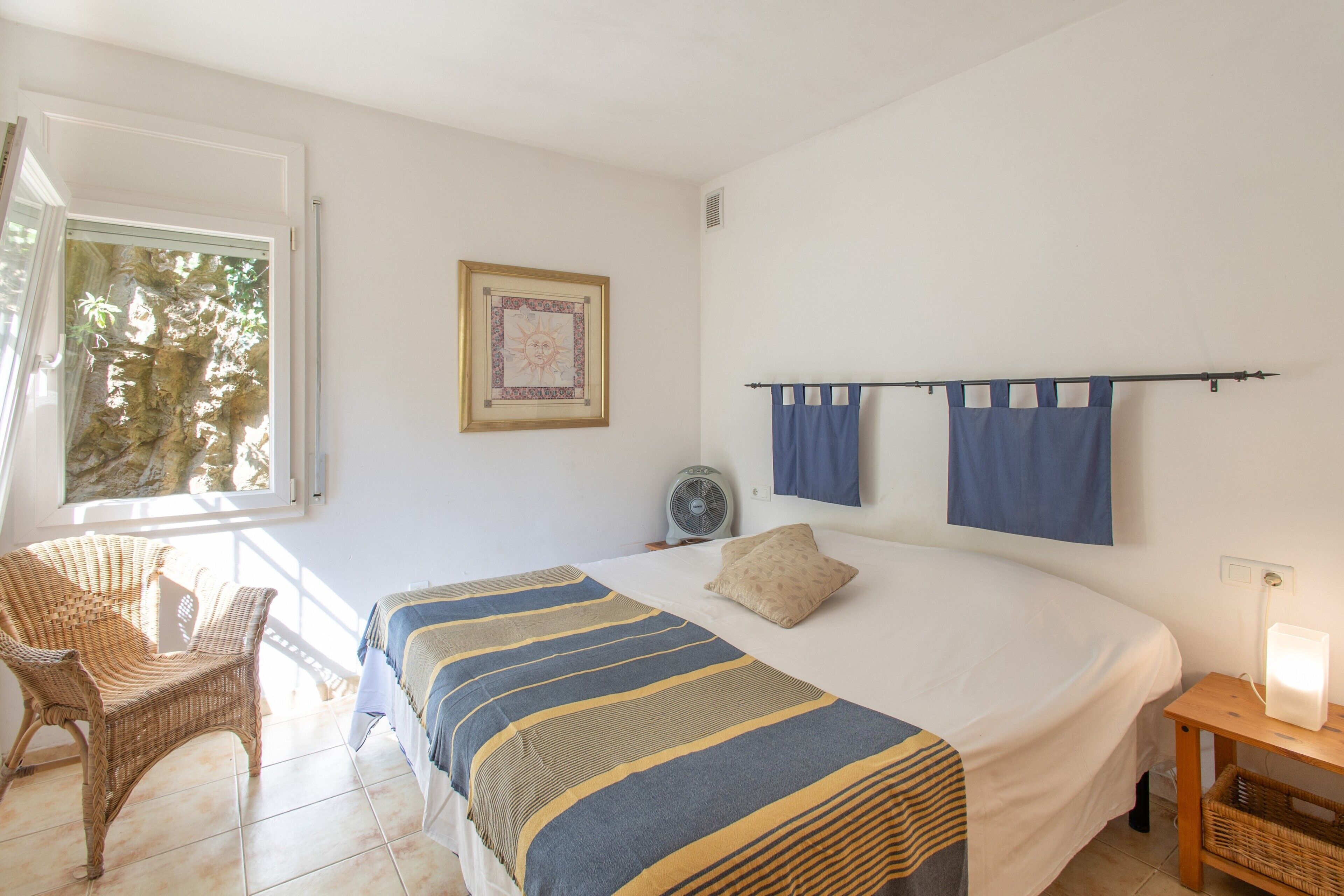 Een slaapkamer met een bed, een stoel, een nachtkastje, een ventilator aan de muur en een raam met gordijnen.