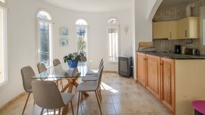 Een eetgedeelte met een glazen tafel en stoelen, een keuken met houten kasten en grote ramen.