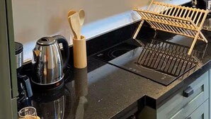 Une cuisine avec un plan de travail noir, une bouilloire, une machine à café, un four à micro-ondes et un étagère en bois.