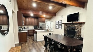 A spacious kitchen with wooden beams, a stone fireplace, and a dining area with a long table and chairs. - 3-bedroom cabin in amazing Running Springs with cozy vibes (Running Springs)
