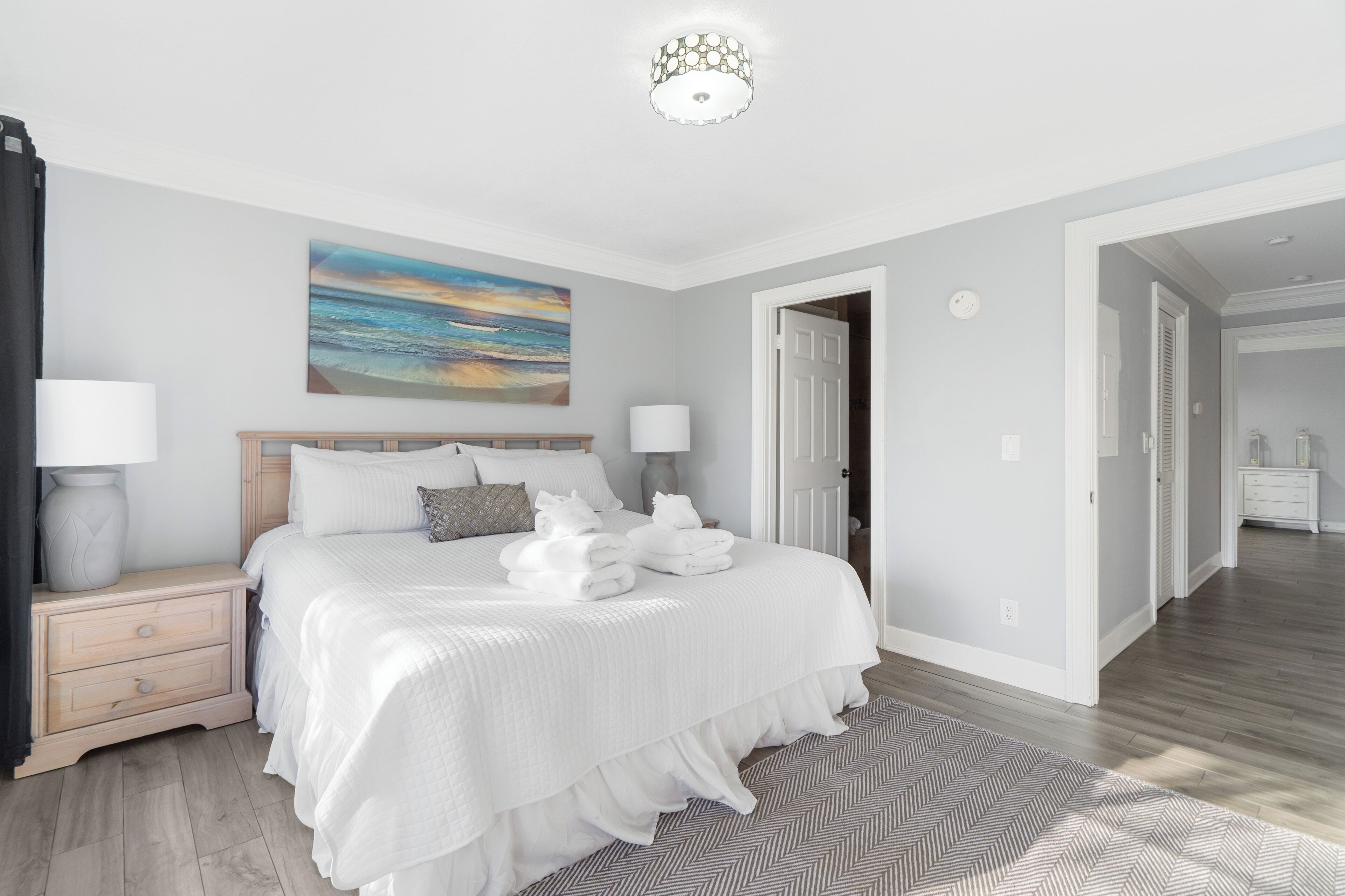 A modern bedroom with a large bed, bedside tables, a painting, and a hallway leading to another room.