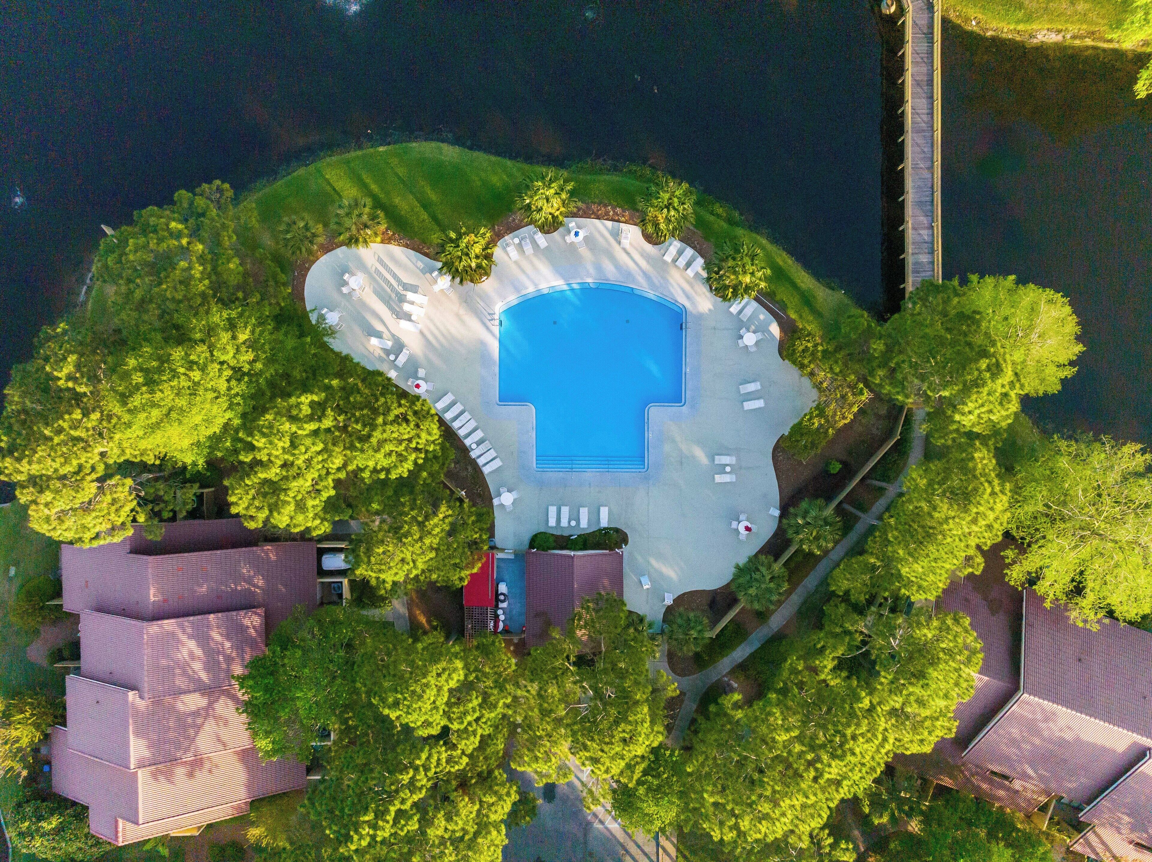 An aerial view of a resort with a central swimming pool, surrounded by lounge areas and trees.