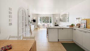 Una cocina moderna con armarios blancos, un comedor con mesa de madera y vistas al patio por la puerta.