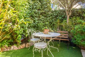 Un jardín con una mesa y sillas de metal, rodeado de abundante follaje y un banco de madera.