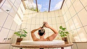 A person in a bathtub with a skylight above, surrounded by tiled walls and potted plants. - Pousada Medeiros  (Monte Verde)