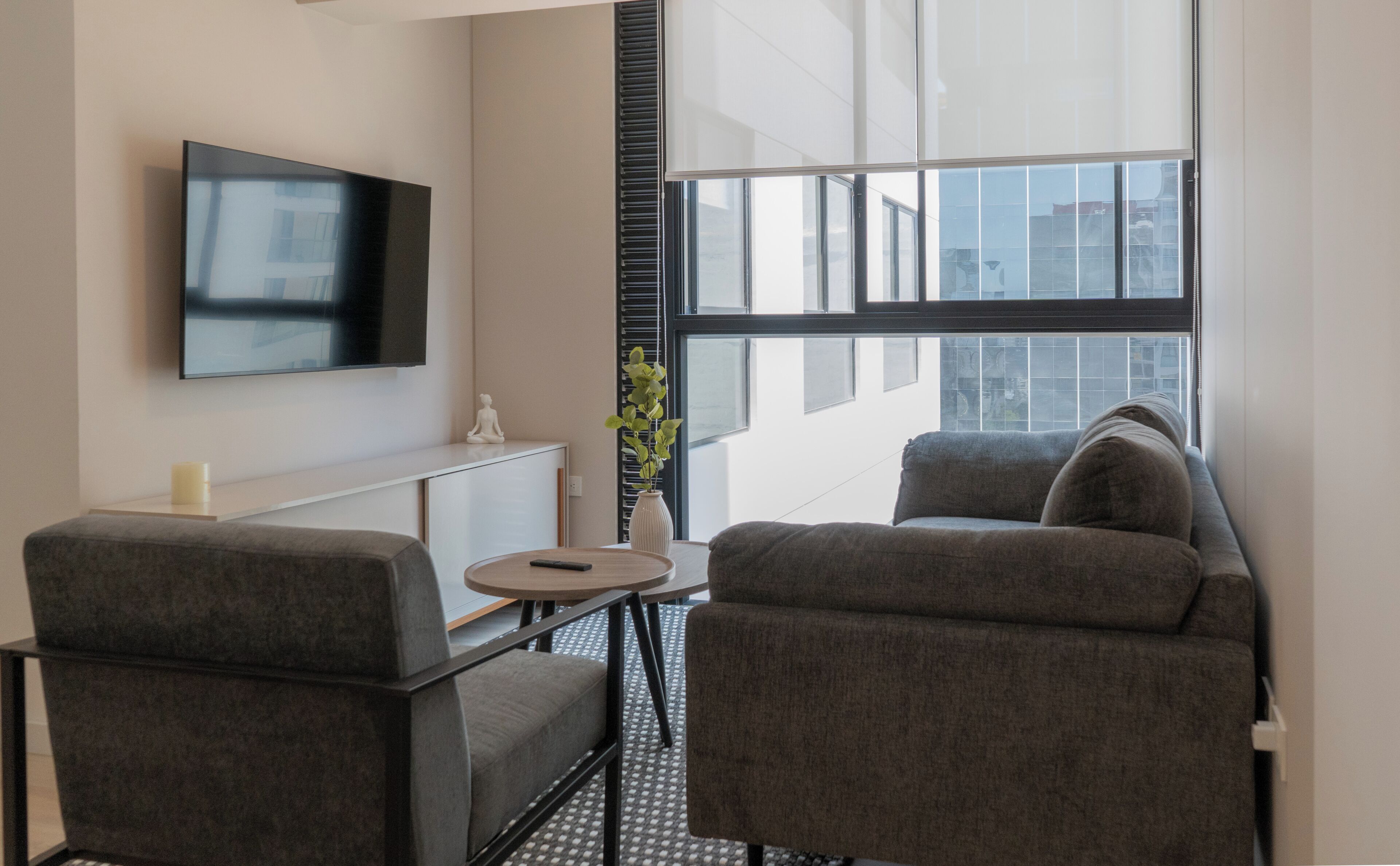 A modern living room with a flat-screen TV, two grey armchairs, a small round table, a vase with a plant, and a window with blinds.