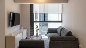 A modern living room with a grey sofa, a small white table, a TV mounted on the wall, and a large window with blinds. - Amme by Wynwood House (Lima)