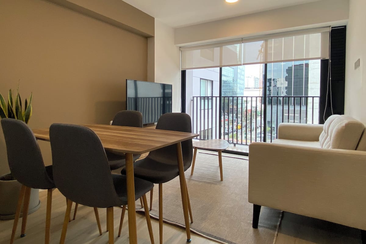 Un salon moderne avec une table à manger en bois, des chaises grises, un canapé beige et une grande porte-fenêtre coulissante donnant sur un balcon.