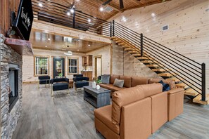 A spacious living area with a leather sofa, a coffee table, and a fireplace. A staircase with a wooden railing leads to an upper level. - Play-Filled Forest Retreat for Large Families (Broken Bow)