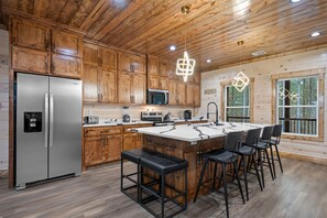 A modern kitchen with wooden cabinets, a stainless steel refrigerator, a built-in microwave, and a large island with bar stools. - Play-Filled Forest Retreat for Large Families (Broken Bow)