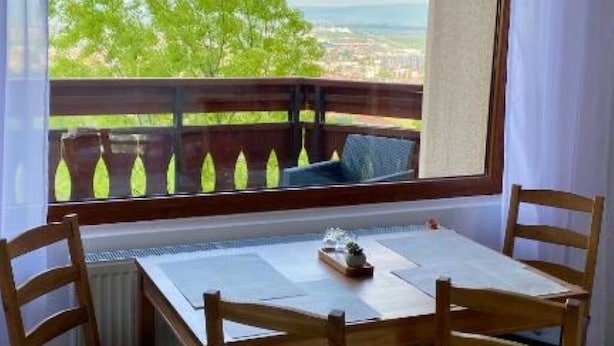 Un balcón con una mesa y sillas, vistas a zonas verdes y edificios, y una ventana con una celosía decorativa.
