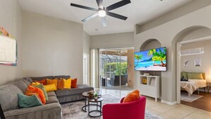 Una sala de estar con un sofá gris, cojines coloridos, un sillón rojo, un televisor de pantalla plana y un ventilador de techo.