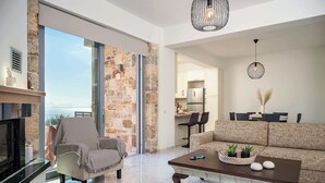 Uma sala de estar ampla com sofá, poltrona e mesa de centro, lareira e vista através de uma porta de vidro.