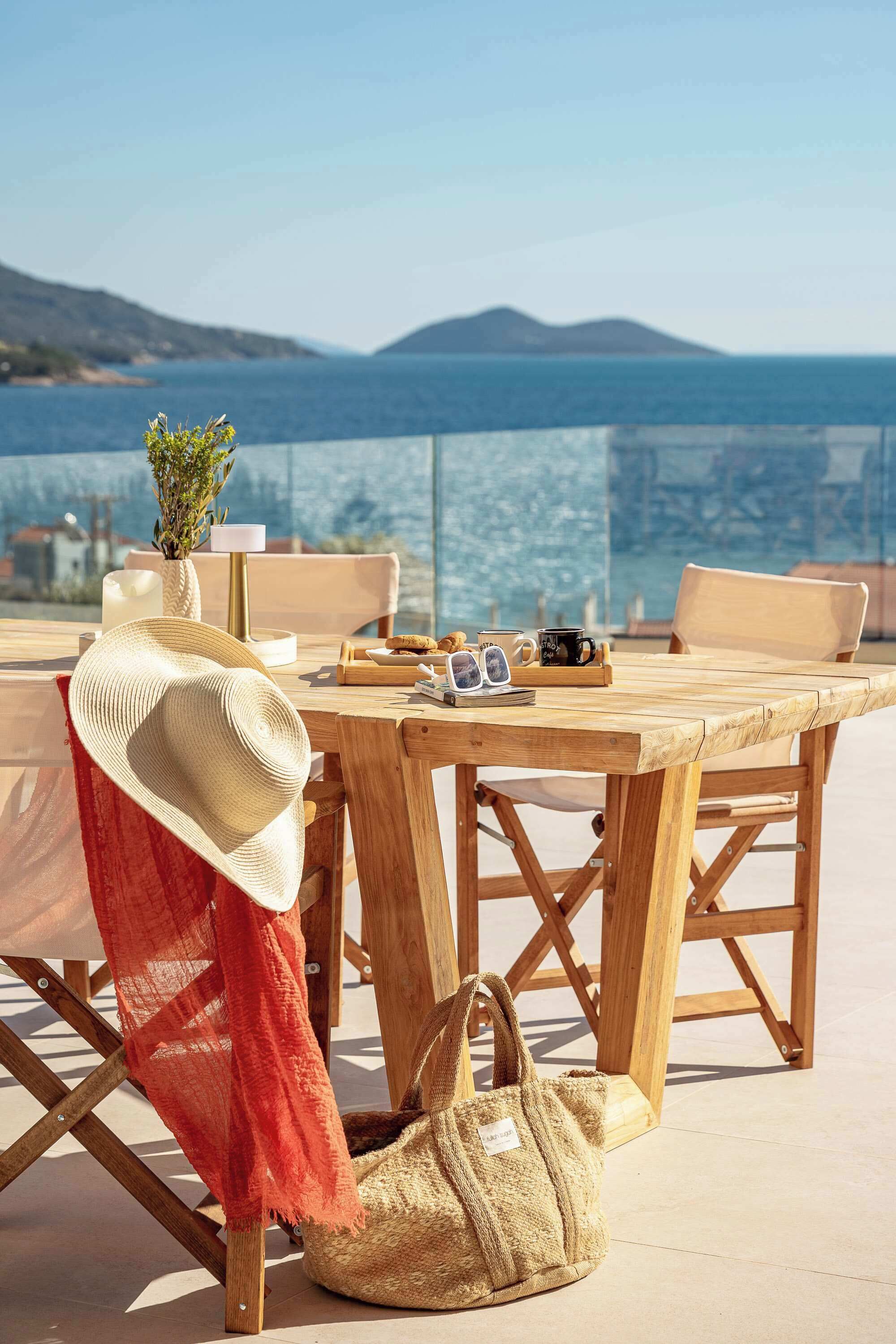 A wooden table with a hat, sunglasses, and a plant on it, set against a scenic view of the sea.