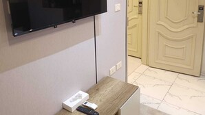Habitación con un televisor de pantalla plana montado en la pared, una mesita consola de madera con un control remoto y una puerta con placa de número.