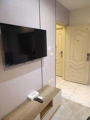 Habitación con un televisor de pantalla plana montado en la pared, una mesita consola de madera con un control remoto y una puerta con placa de número.