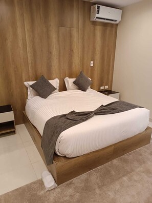 A bedroom with a wooden headboard, a bed with white bedding and two pillows, a bedside table, and an air conditioning unit. - Tamaya Hotel Fifth Settlement (Shurtah al-Dakhlah)
