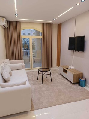 A modern living room with a sofa, a small table, a TV mounted on the wall, and a sliding glass door leading to an outdoor area. - Tamaya Hotel Fifth Settlement (Shurtah al-Dakhlah)