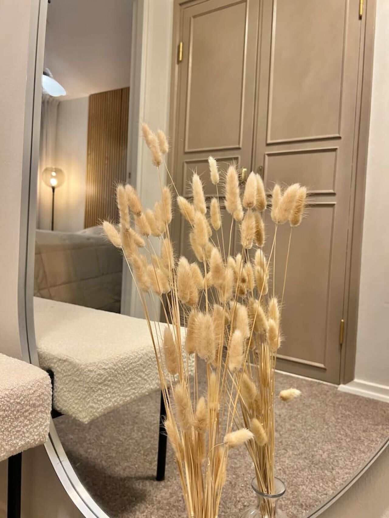 A room with a beige sofa, a glass coffee table, and a tall vase with dried pampas grass.