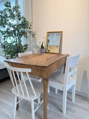 Una mesa de comedor con tapa de madera, rodeada de sillas blancas, un espejo con marco dorado y una planta en maceta.