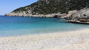 Ein Kiesstrand mit klarem Wasser, ein felsiger Hang und ein Steg, der ins Meer führt.