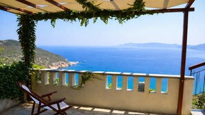 Eine Steinterrasse mit einem Holzstuhl, einer mit Wein bewachsenen Pergola und Blick auf das Meer und die entfernten Hügel.