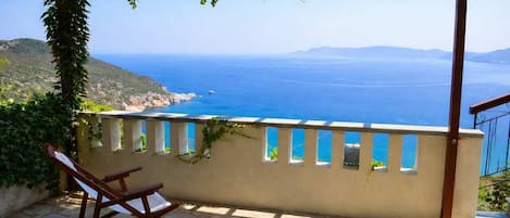 A stone patio with a wooden chair, a pergola with vines, and a view of the sea and distant hills.