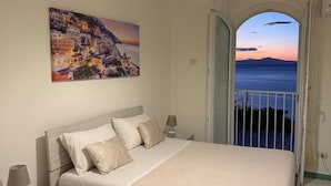 Quarto com cama, mesas de cabeceira, luminária, pintura e vista para o mar por uma porta aberta.