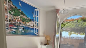 Uma sala de estar com sofá, um quadro de uma cidade costeira, um abajur e vista para uma varanda com vegetação.