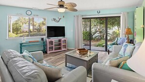 A living room with a sofa, coffee table, TV, and a balcony with curtains. - Crystal Village II 26A (Miramar Beach)