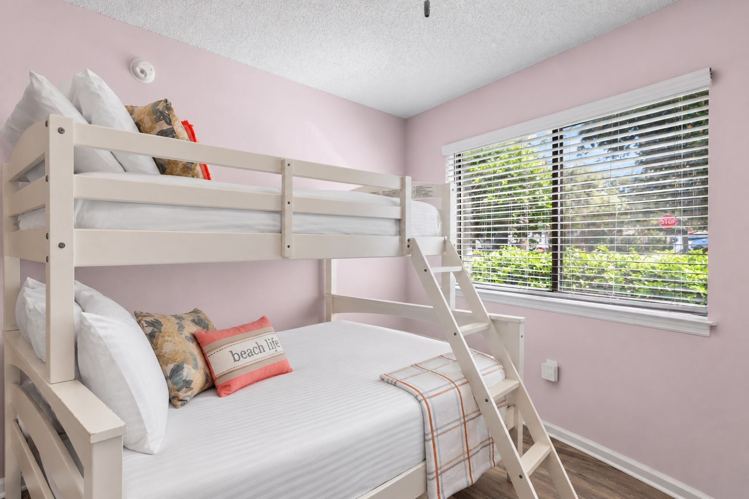 Habitación con literas, ventana con persianas, escalera y almohada con la frase 'beach life'.