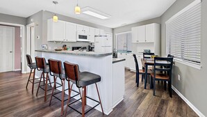 A modern kitchen with white cabinets, a central island with bar stools, and a dining area with a table and chairs. - Crystal Village II 26A (Miramar Beach)