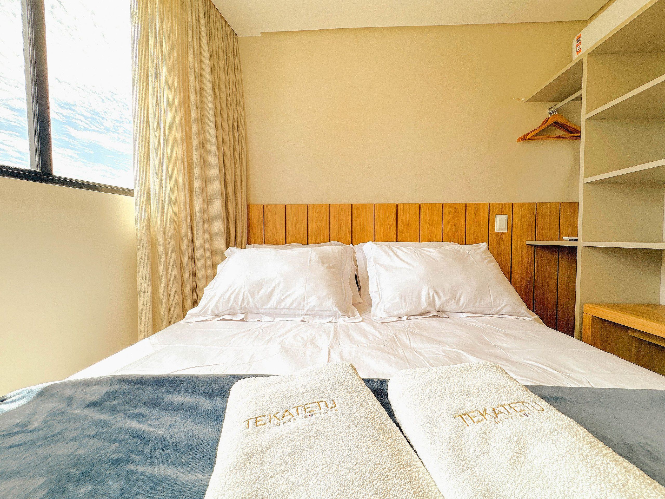 A hotel room with a bed, two towels with 'TEKATETU' branding, a wooden headboard, a wardrobe, and a window with curtains.