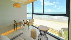 Ein Zimmer mit großem Fenster mit Blick auf den Strand, einem runden Tisch, zwei Stühlen, einer Couch und einer Pendelleuchte.