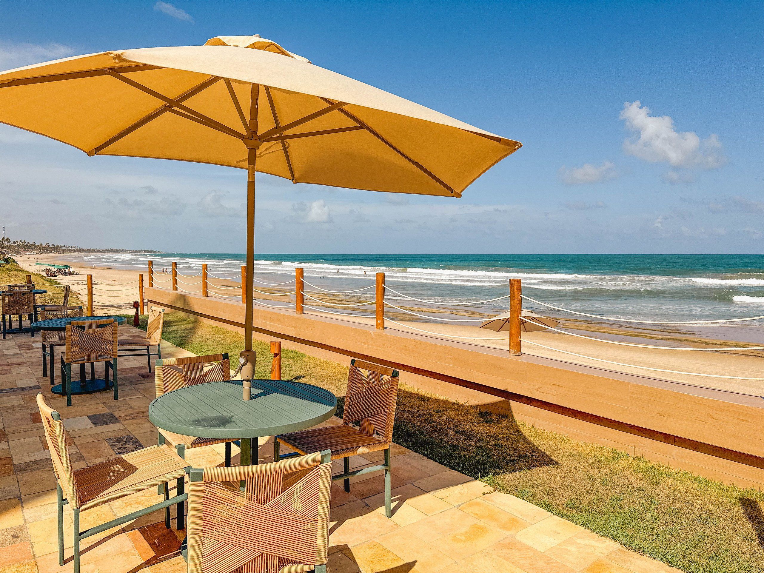 Un patio frente a la playa con mesas y sillas, una sombrilla grande y vista al océano.