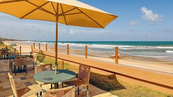 Un patio frente a la playa con mesas y sillas, una sombrilla grande y vista al océano.