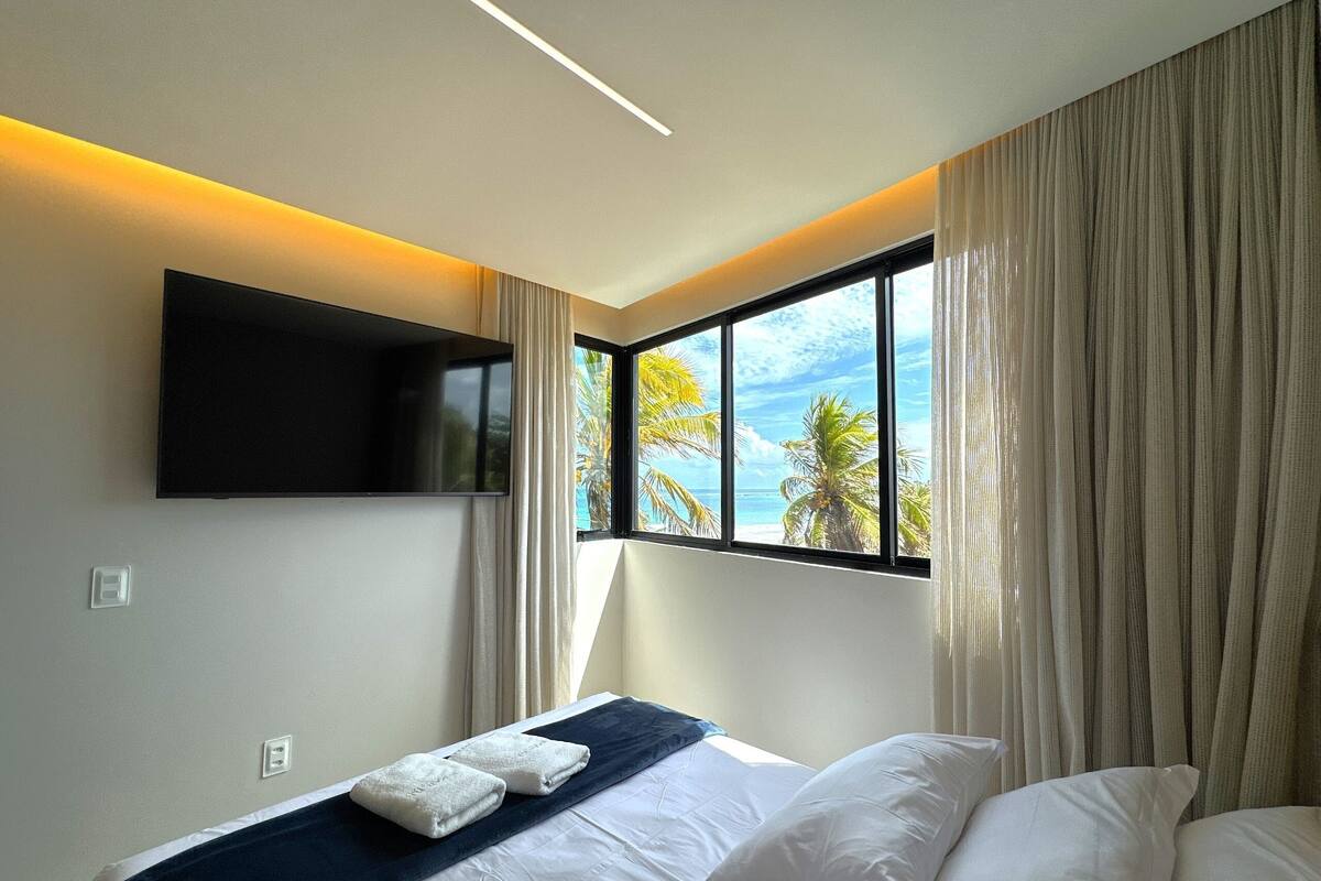 Ein Hotelzimmer mit Bett, einem an der Wand montierten Fernseher, einem Fenster mit Vorhängen und Blick auf Palmen und das Meer.