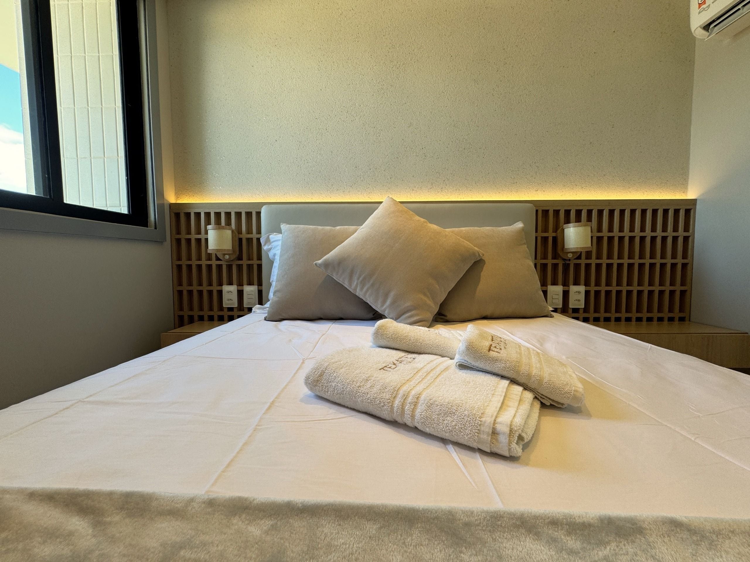 A neatly made bed with pillows, a towel, and a headboard with wall-mounted lights.