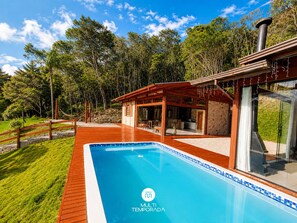 A modern outdoor pool area with a wooden deck, a glass-enclosed structure, and a clear blue sky. - Chácara Pôr-do-Sol Sto Amaro Imperatriz (Santo Amaro da Imperatriz)