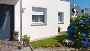 Una casa moderna con fachada blanca, puerta principal, un pequeño porche y un jardín con hortensias y otras flores.