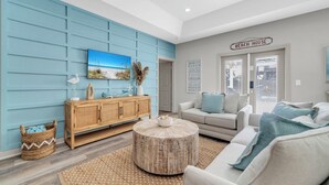 A living room with a blue wall, a wooden cabinet, a round coffee table, a sofa, and a sign reading 'BEACH HOUSE'. - 🏖️Walk to Beach | Great Location | Fire Pit | Sandy Shores🏖️ (Panama City Beach)