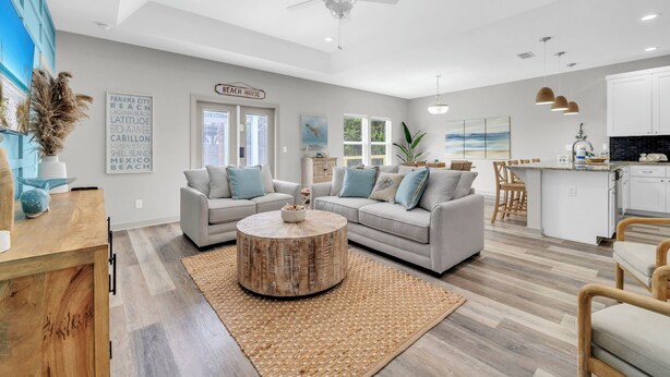 A spacious living area with a large round coffee table, two sofas, a ceiling fan, and a kitchen area with white cabinets. - 🏖️Walk to Beach | Great Location | Fire Pit | Sandy Shores🏖️ (Panama City Beach)