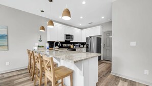 A modern kitchen with white cabinets, a granite countertop, stainless steel appliances, and wooden bar stools. - 🏖️Walk to Beach | Great Location | Fire Pit | Sandy Shores🏖️ (Panama City Beach)