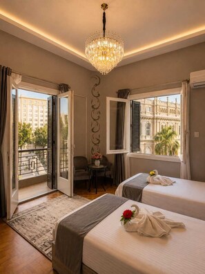 A hotel room with two beds, a chandelier, a balcony with a view of buildings, and a small table with chairs. - Garden City Tahrir Downtown (Cairo)