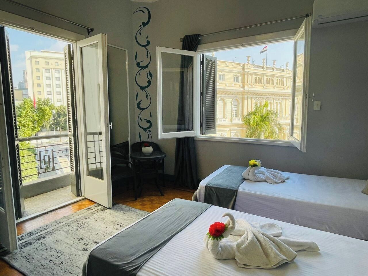 Habitación de hotel con dos camas, un balcón, vistas a un edificio y una pequeña mesa con una silla.