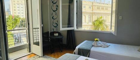 Habitación de hotel con dos camas, un balcón, vistas a un edificio y una pequeña mesa con una silla.