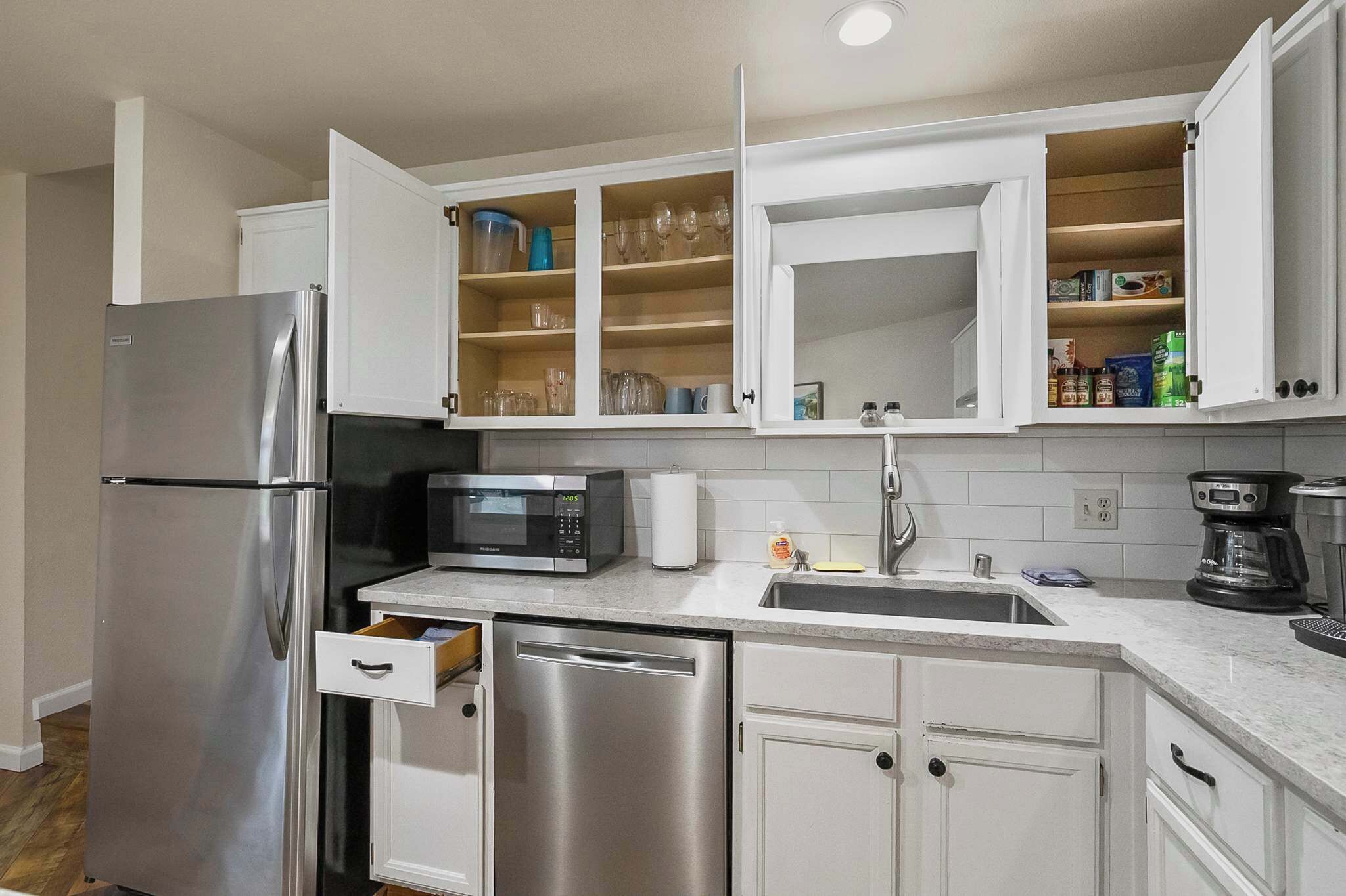 A modern kitchen with a stainless steel refrigerator, microwave, and dishwasher, white cabinetry, and a countertop with a coffee maker and sink.