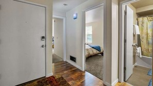 En hall med trägolv, en dörr, en ljusströmbrytare och en säng med ett blått täcke i bakgrunden.