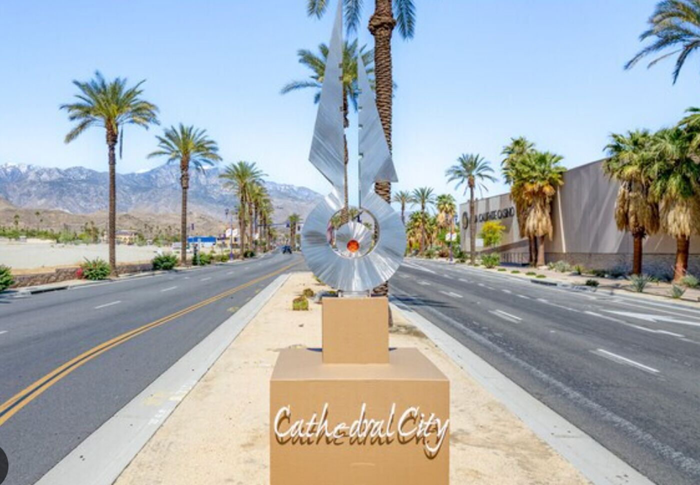 Una carretera bordeada de palmeras, una gran escultura metálica y un cartel que dice 'Cathedral City'.