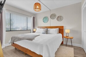 A bedroom with a wooden bed, two bedside lamps, a window with blinds, and three wall decorations. - Beachfront Pet-Friendly 2BR Condo Walkable to Boardwalk (Carolina Beach)